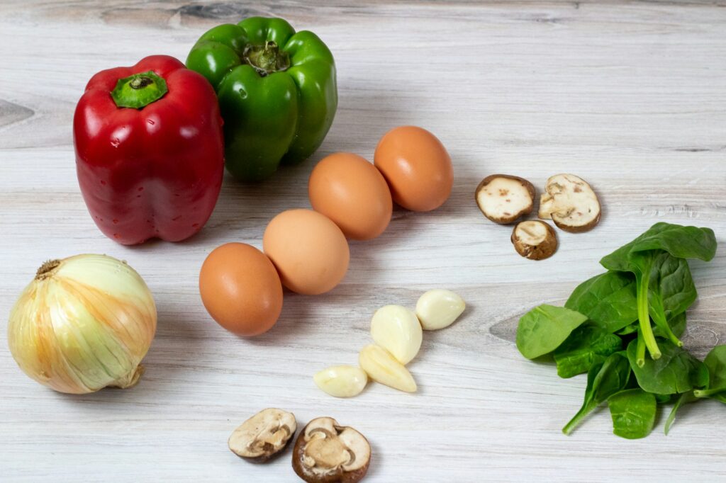 Red and green bell peppers, an onion, five brown eggs, sliced mushrooms, garlic cloves and fresh spinach on a white wooden table