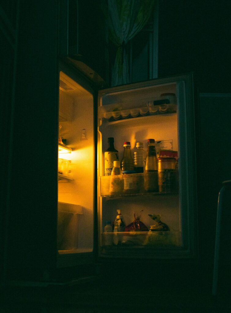 An open refrigerator glowing in a dark kitchen, bottles and jars on its door and shelves