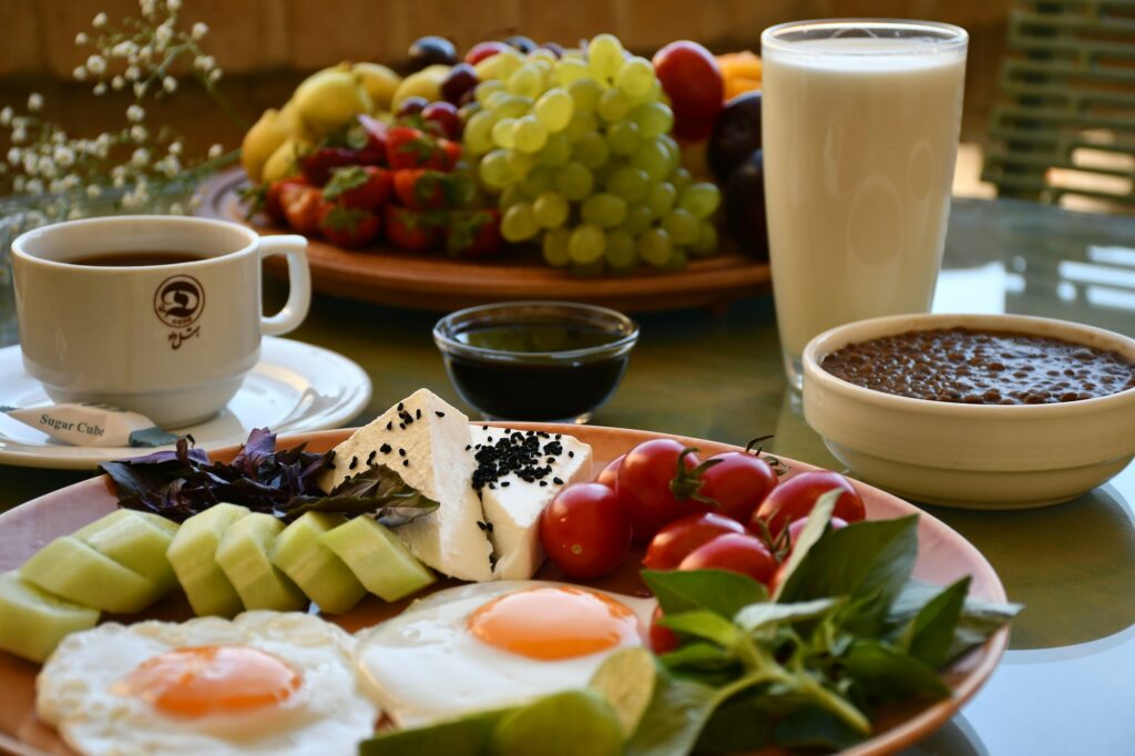 A breakfast table with two fried eggs, cucumber, tomatoes and feta, a cup of coffee, a glass of milk and a platter of fruit