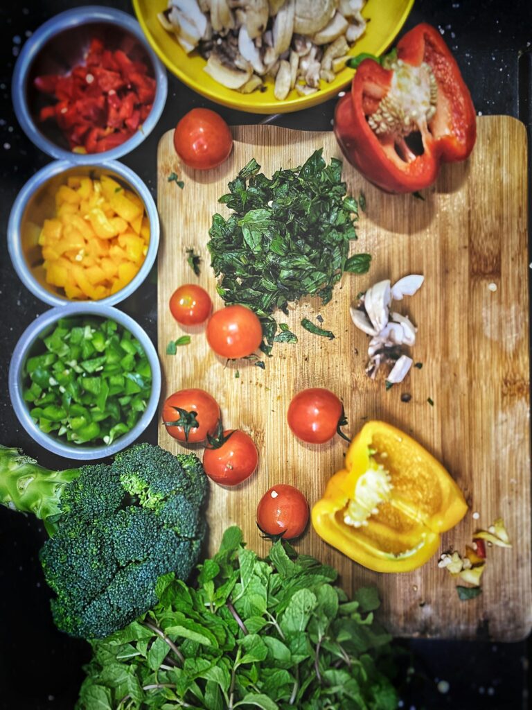 Bowls of chopped red, yellow and green peppers and sliced mushrooms beside a cutting board with tomatoes and herbs