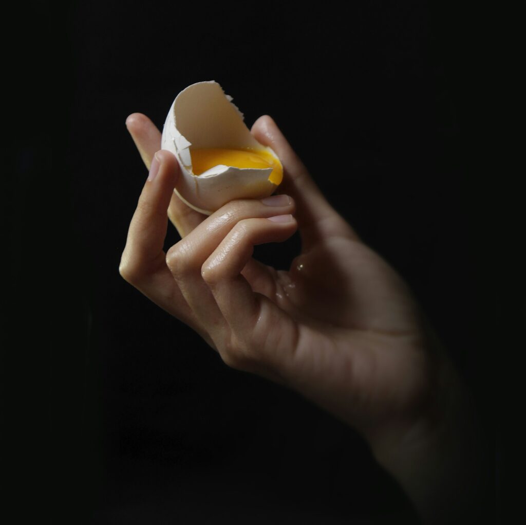 A hand holding half of a cracked egg shell with the yolk inside against a black background