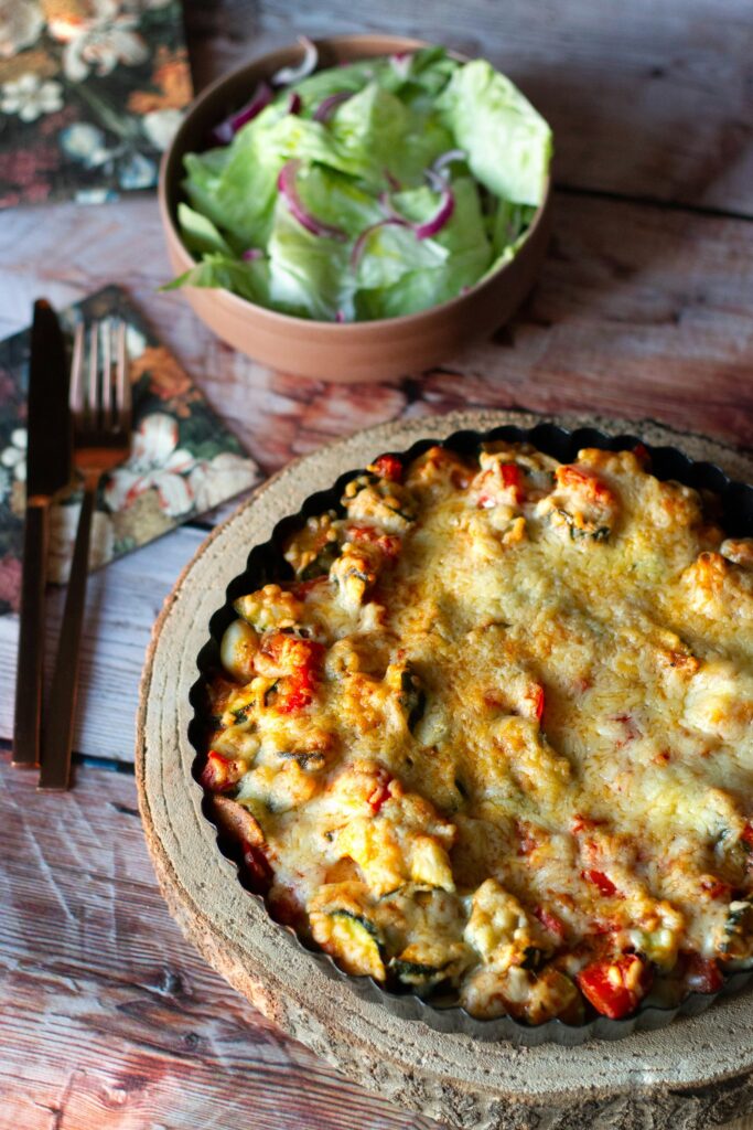 A vegetable bake with red peppers and zucchini under a thick layer of melted cheese, a green salad behind